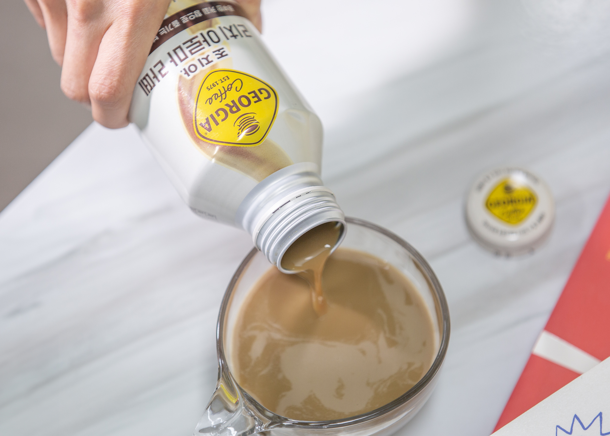 테이블 위에 투명한 유리잔이 놓여 있고, 누군가가 조지아 리치 아로마 라떼 제품을 손에 들고 기울여서 컵에 가득 따르고 있다.