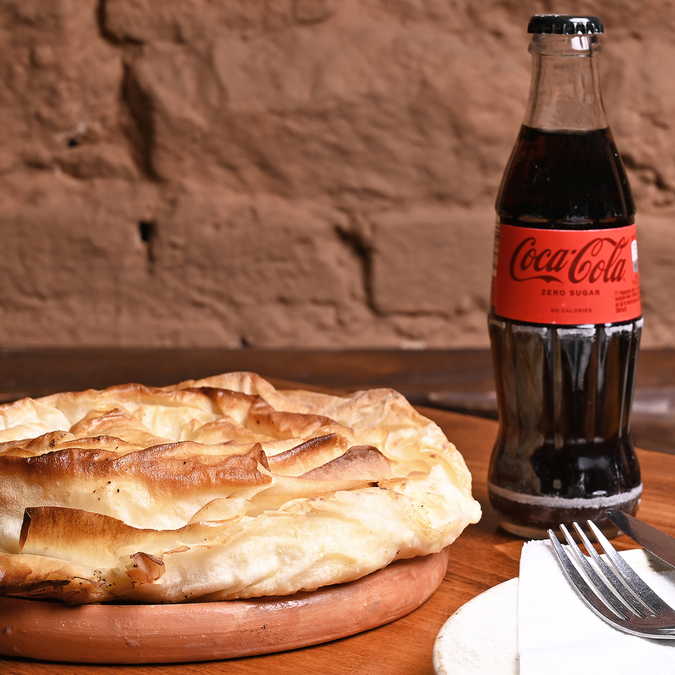 Tavë me pite të pjekur dhe shishe Coca-Cola Zero në një tavolinë druri.