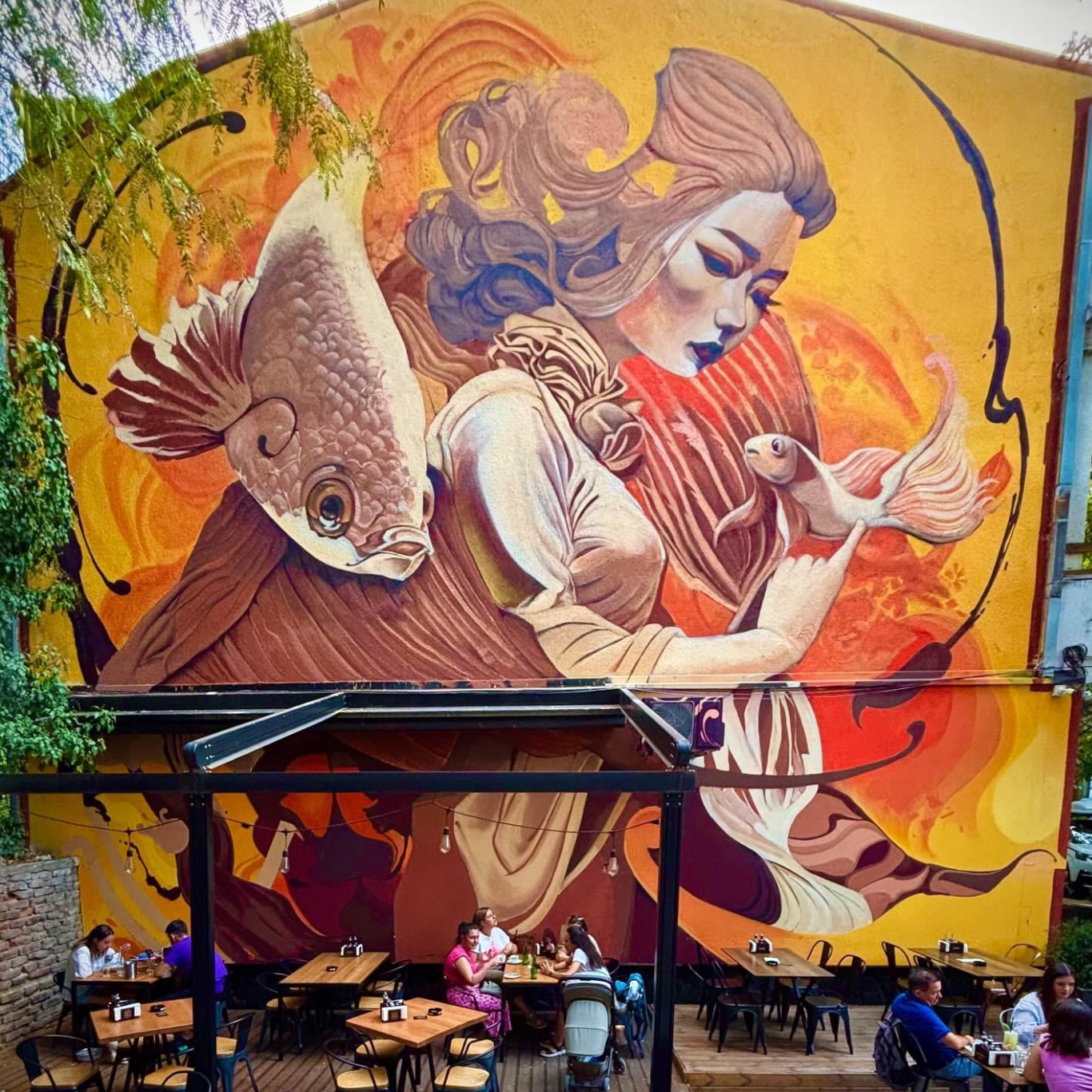 Mural i madh me një grua dhe peshq mbi një tarracë me njerëz që ulen dhe shijojnë.