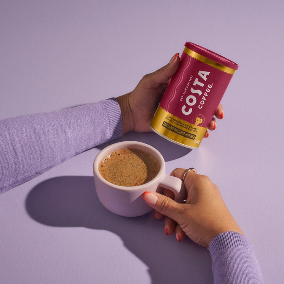 Una persona agarrando una taza de café y con la otra mano sosteniendo una lata de café soluble Costa Coffee.
