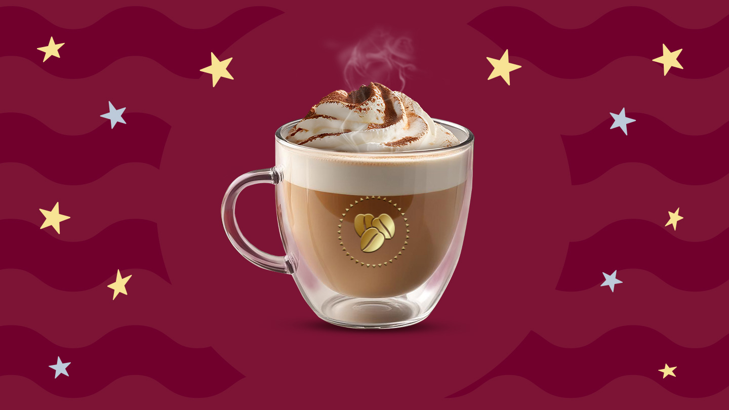 Una taza de cristal con cinnamon latte caliente y crema batida por encima decorado con canela en polvo por encima sobre un fondo en tonos guinda.