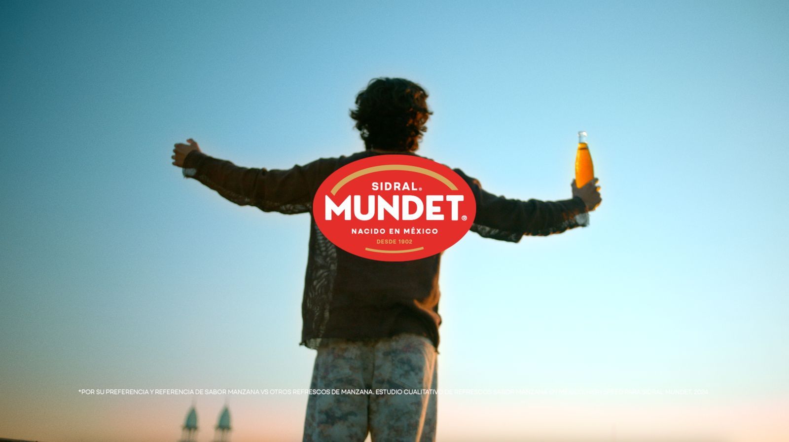 Hombre de espaldas con los brazos extendidos, sosteniendo una botella de Mundet al atardecer. El logo de Mundet se superpone en la imagen.