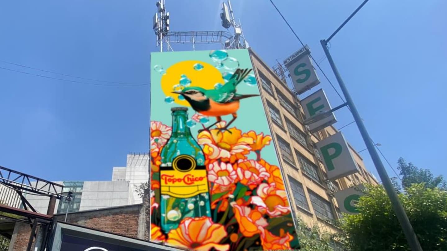 Un mural en un edificio muestra una botella de Topo Chico, un pájaro colorido y flores naranjas, todo bajo un sol amarillo. El cielo azul es visible en la parte superior.
