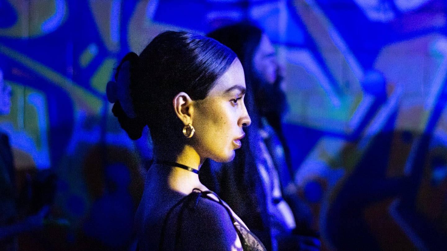 Una mujer con el pelo oscuro recogido se ve de perfil contra una pared azul cubierta de grafiti. Lleva un collar ajustado y pequeños pendientes.