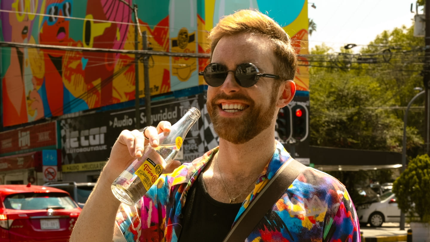 Un hombre con barba y gafas de sol sonríe mientras sostiene una botella de Topo Chico frente a un mural colorido en México. Un coche rojo y árboles son visibles en el fondo.