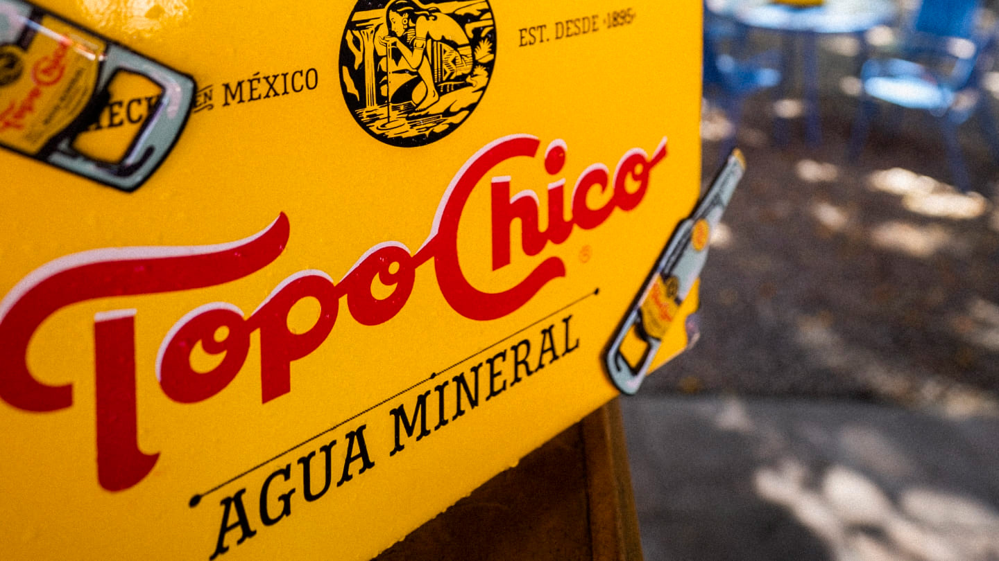 Una hielera amarilla de Topo Chico con el logo rojo brillante y la inscripción "Agua Mineral", con sillas azules borrosas al fondo, sugiriendo un ambiente relajado al aire libre.