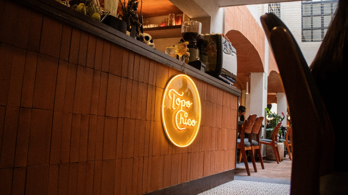 Una vista interior de un elegante bar o restaurante con un letrero de neón de Topo Chico en la pared de ladrillo, con asientos y decoración que sugieren un ambiente moderno y acogedor.