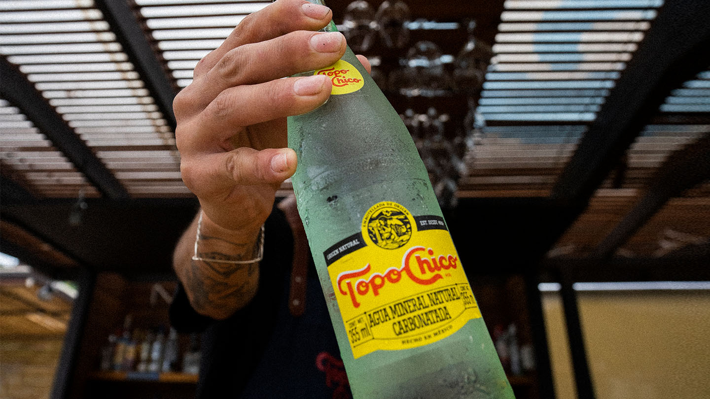 Una mano sostiene una botella helada de Topo Chico con la etiqueta amarilla distintiva, con un fondo borroso que sugiere un bar o restaurante al aire libre.