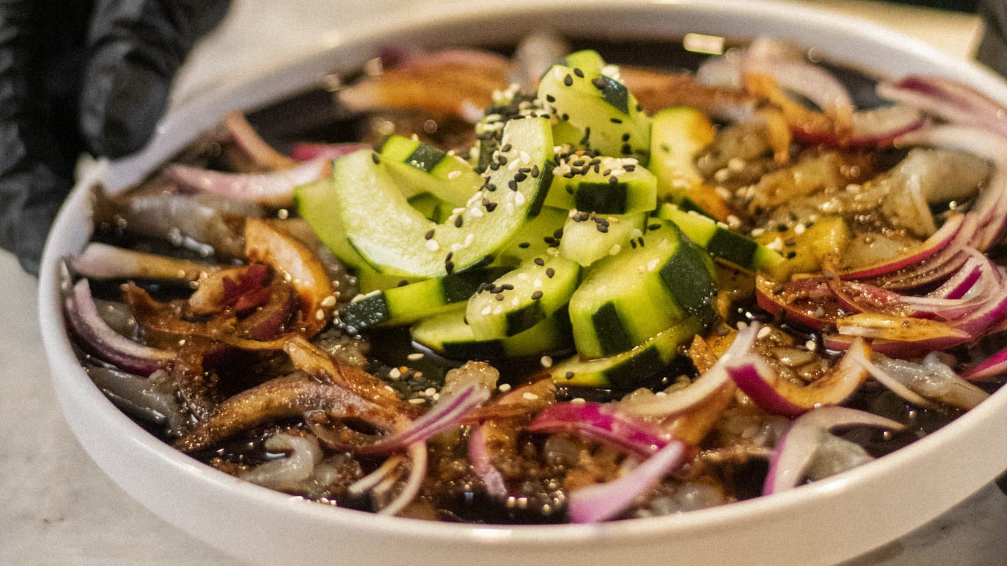 Un plato de comida con una variedad de ingredientes, incluyendo pepino en rodajas, cebolla roja y semillas de sésamo, presentado en un cuenco blanco.