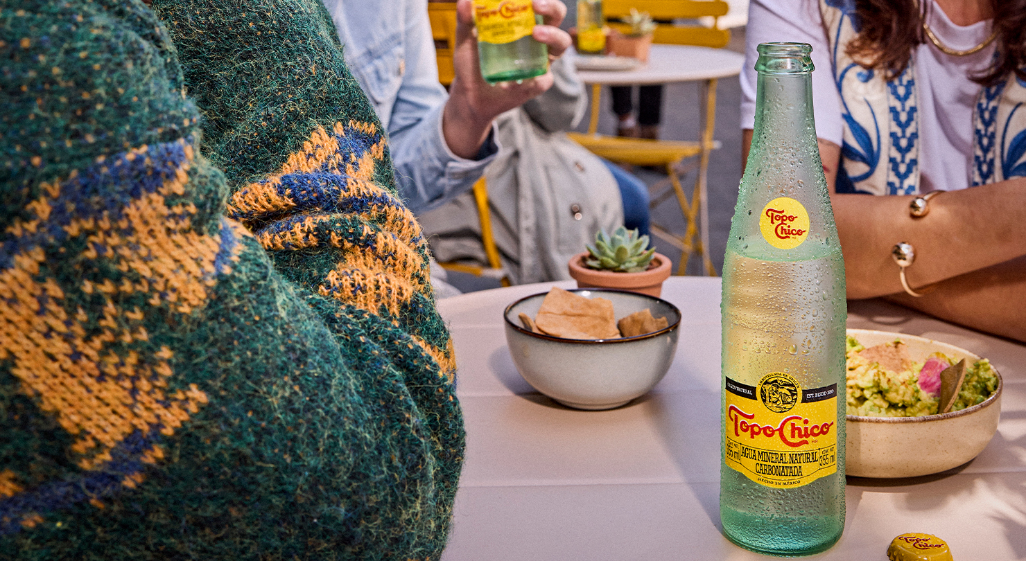 Tres amigos disfrutan de comida y Topo Chico en un café al aire libre.