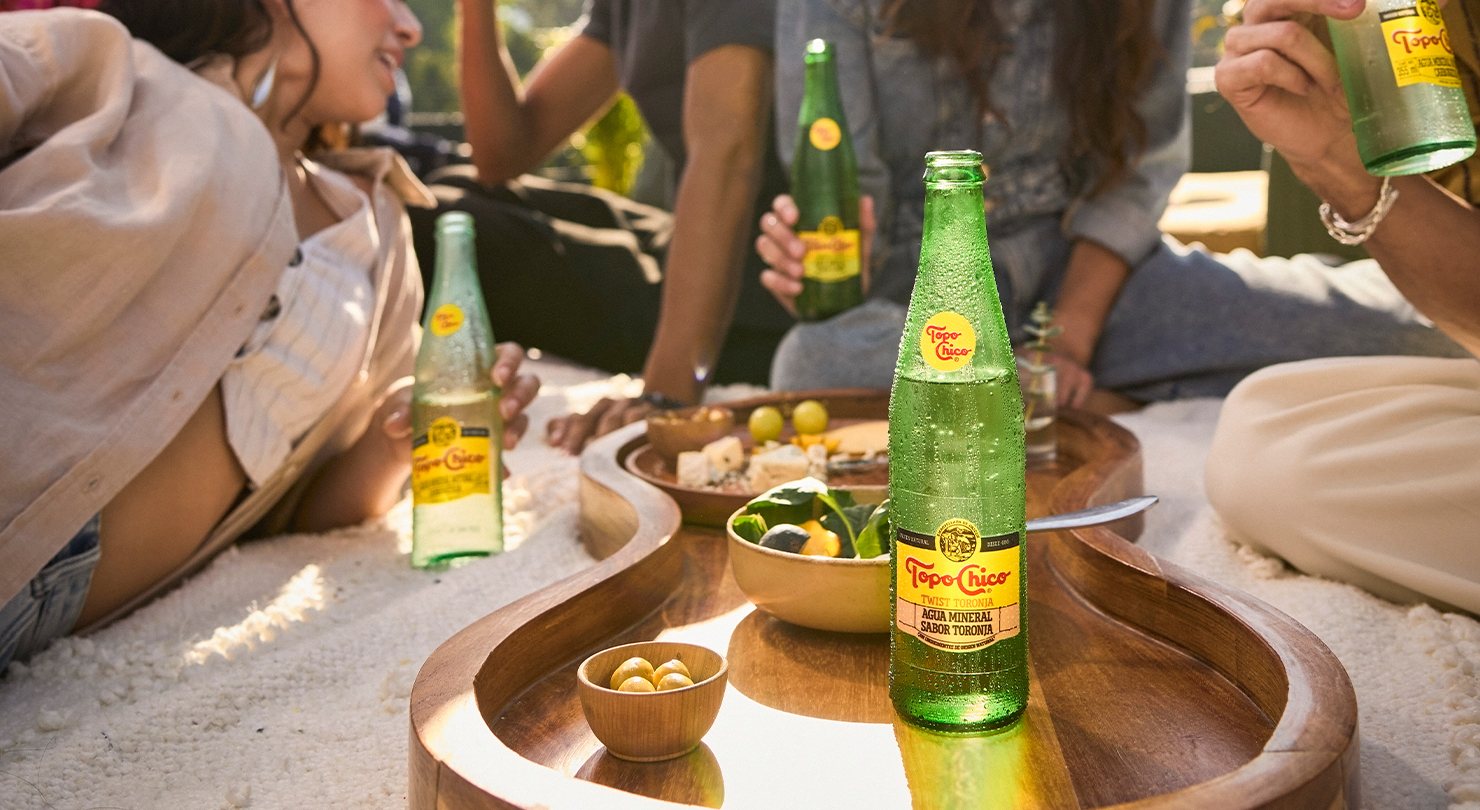 Grupo de amigos de picnic al aire libre, con botellas de Topo Chico y un plato de comida. Están relajados y conversando, con el enfoque en las botellas y la comida en primer plano.