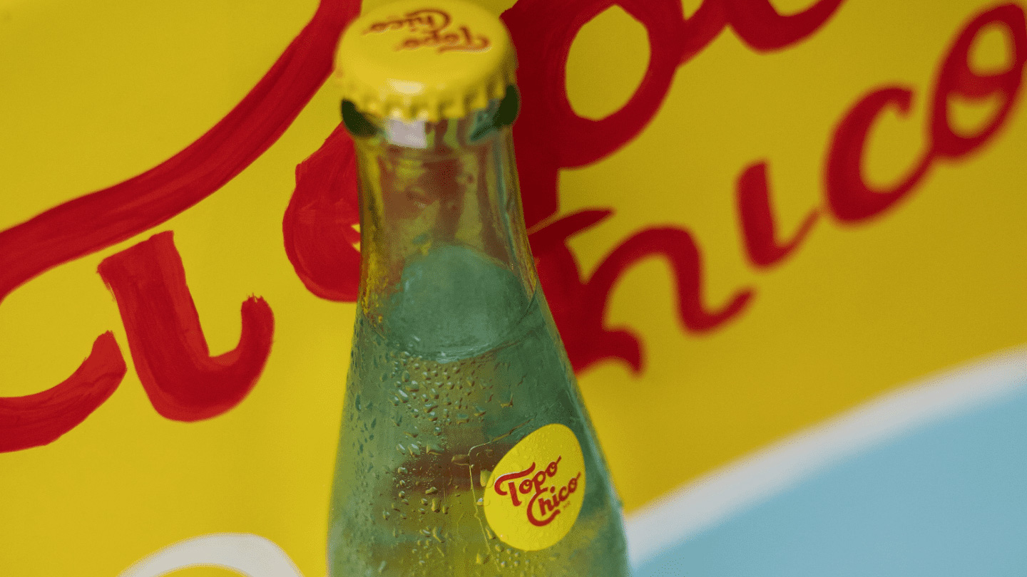 Botella de vidrio Topo Chico. Detrás hay una heladera amarillo Topo Chico