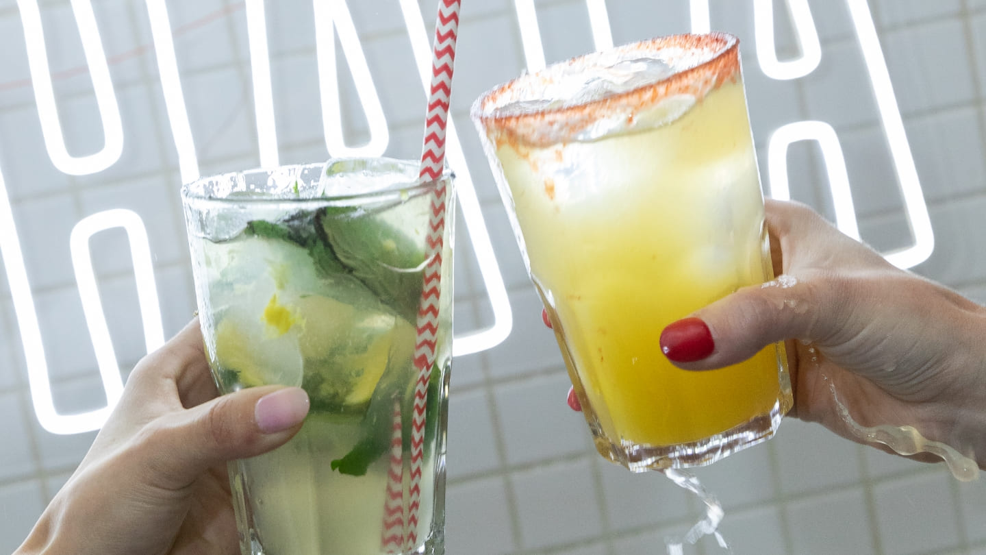 Primer plano de dos manos de mujeres. Cada una sostiene un trago, sobre un fondo con letras de neón.