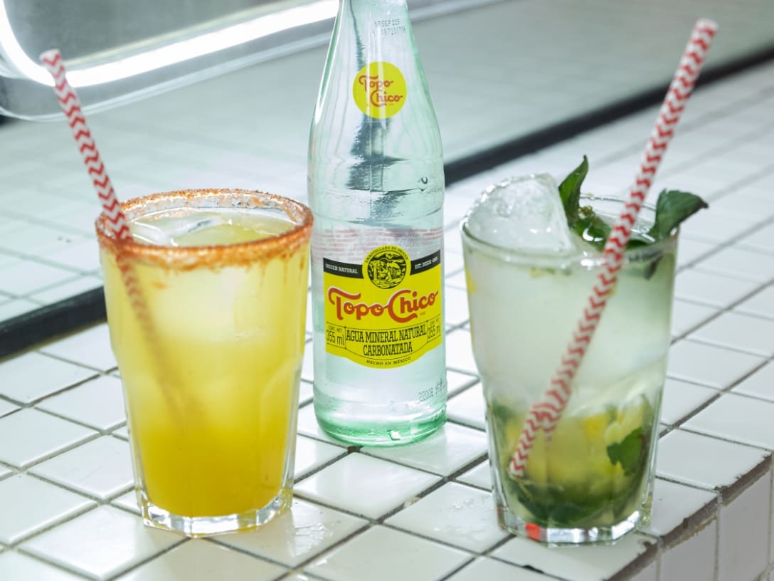 Dos tragos y una botella de Topo Chico sobre una mesa de mosaicos blancos.