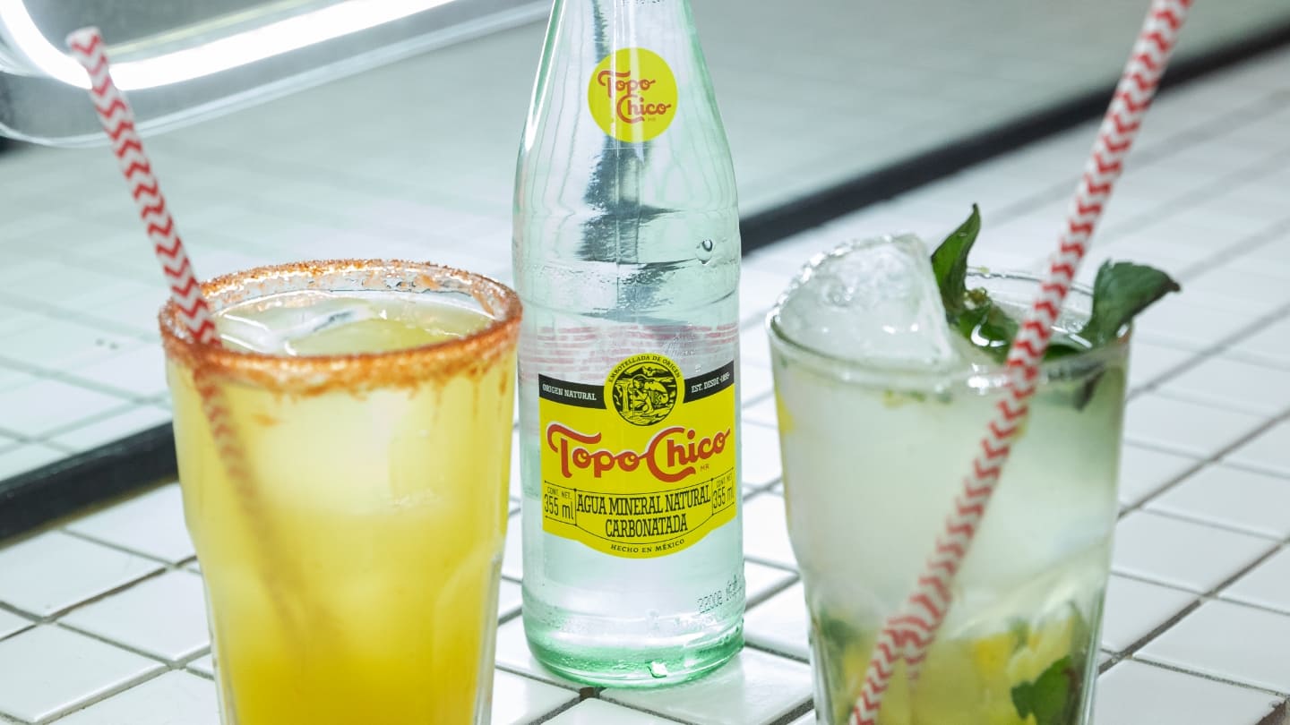 Dos tragos y una botella de Topo Chico sobre una mesa de mosaicos blancos.