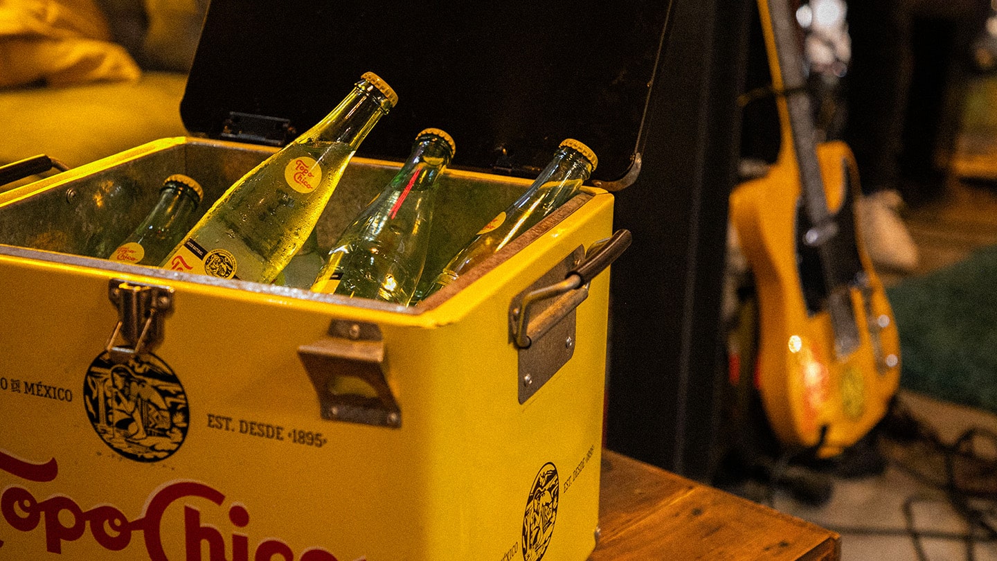 Un refrigerador amarilla con el logo de Topo Chico y dentro hay botellas de vidrio de Agua Mineral Topo Chico en hielo. Detrás se ve una guitarra eléctrica naranja.
