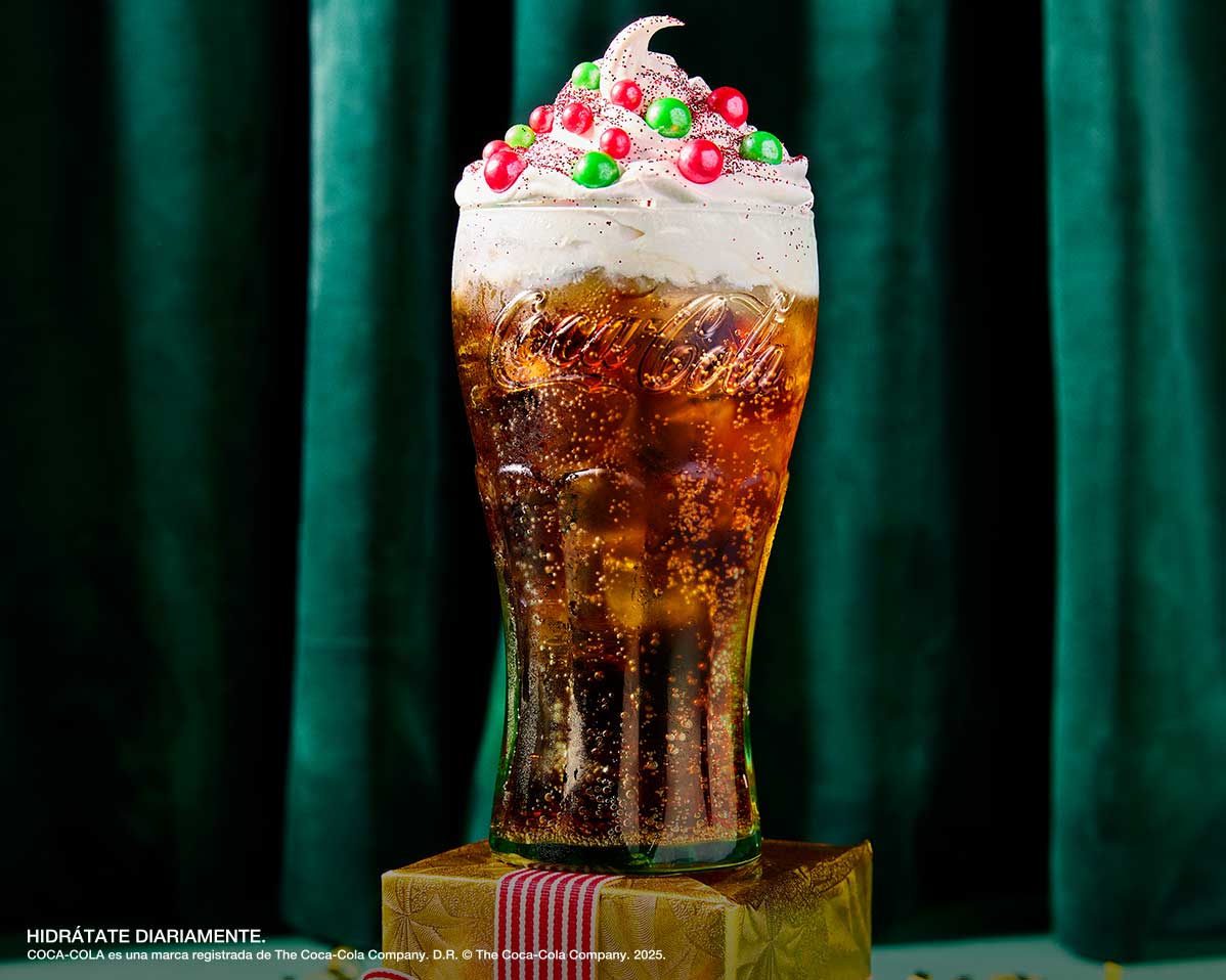 Bebida especial de Coca-Cola con crema batida y decoraciones de colores, presentada en vaso de vidrio sobre base decorativa.
