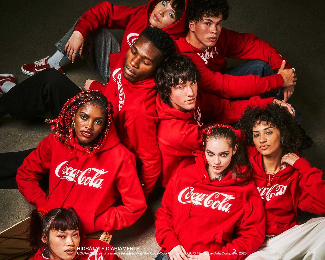 Grupo diverso de jóvenes usando sudaderas rojas de Coca-Cola, sentados juntos en una sesión fotográfica de estilo urbano.​