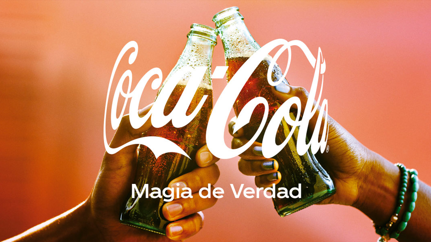 Dos manos que brindan con botellas deCoca-Cola. Encima de la imágen, el logo de Coca-Cola y la frase "Magia de verdad". 
