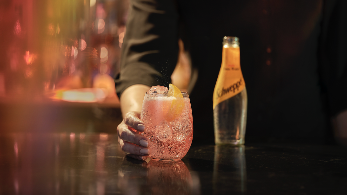 Ruka drži čašu sa roze koktelom i kriškom narandže, dok se na stolu pored nalazi boca Schweppes-a, uz promotivni tekst o novim ukusima i koktelima