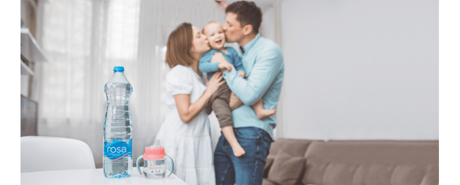 Flaša Rosa vode i dečija čaša sa ručkama na stolu, dok dvoje odraslih drže dete u naručju u pozadini