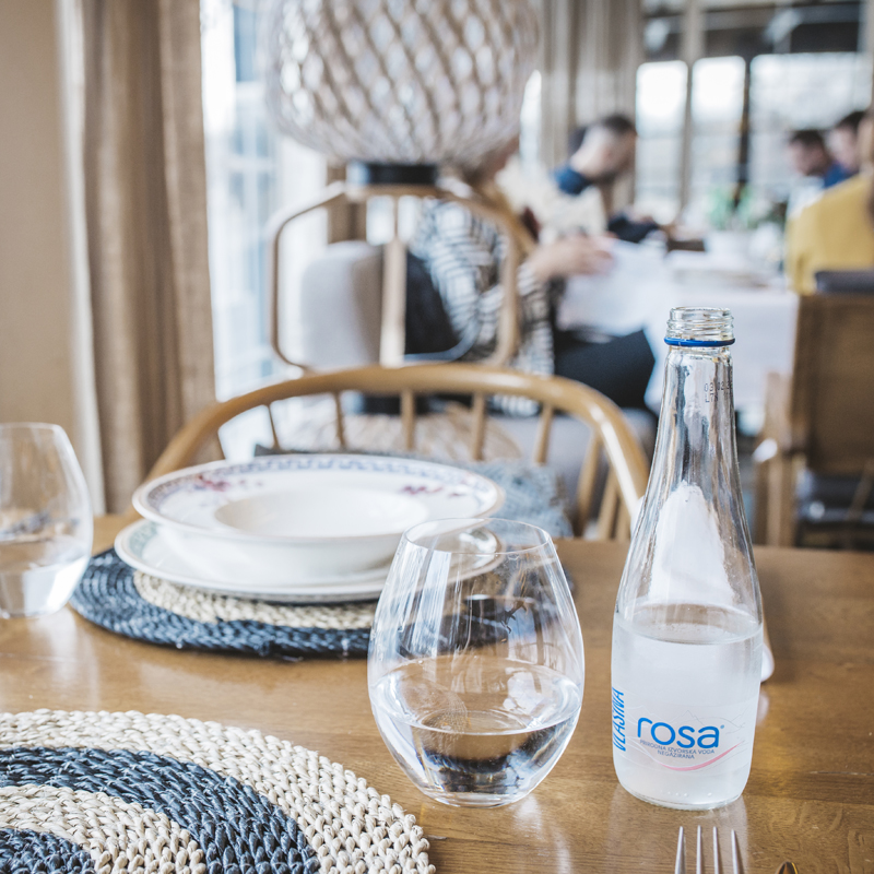 Flaša Rosa vode i čaša na drvenom stolu u restoranu, uz postavljene tanjire i pletene podmetače