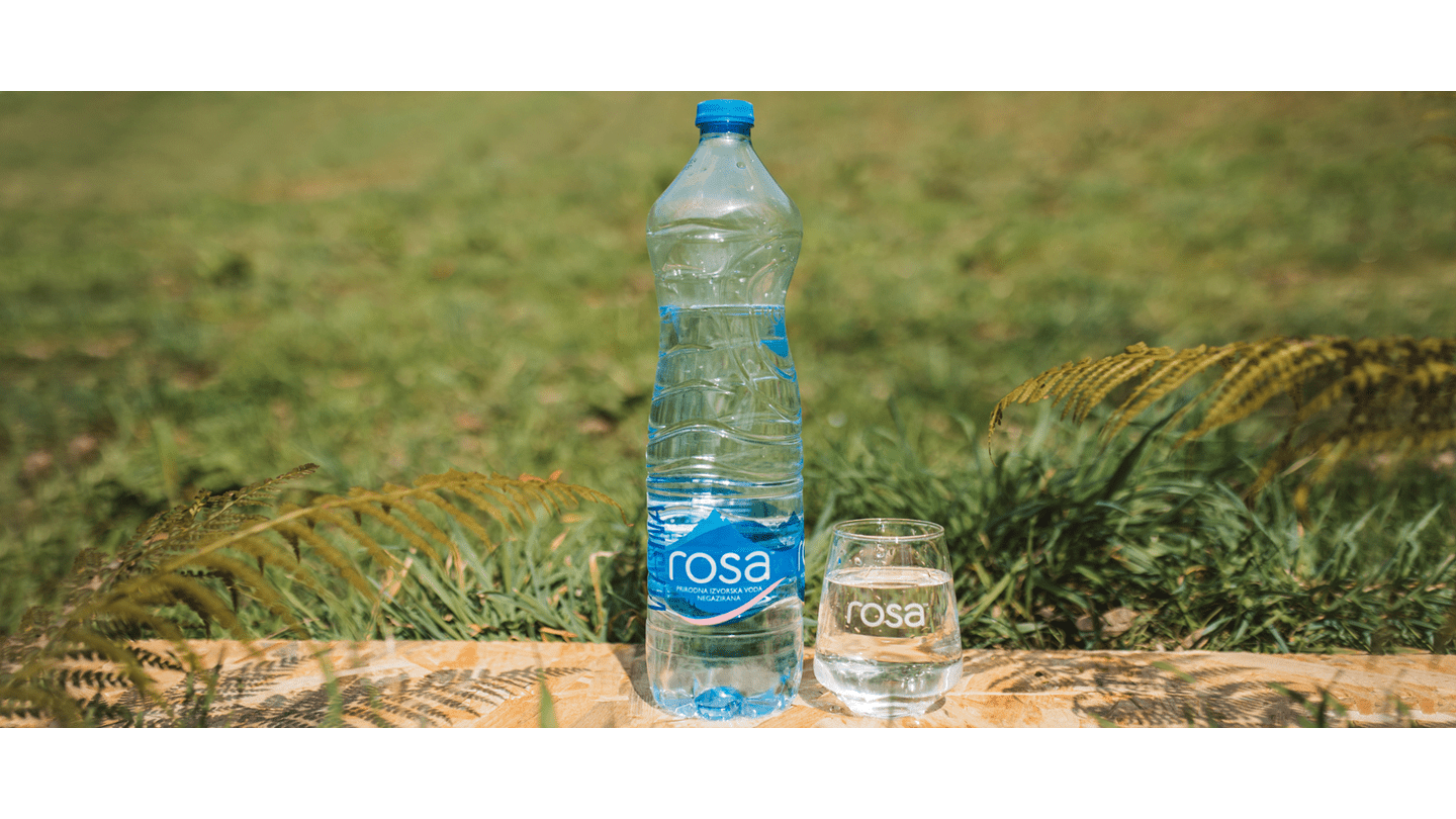 Plastična boca i čaša Rosa negazirane izvorske vode postavljene na drvenu površinu na travnatoj pozadini
