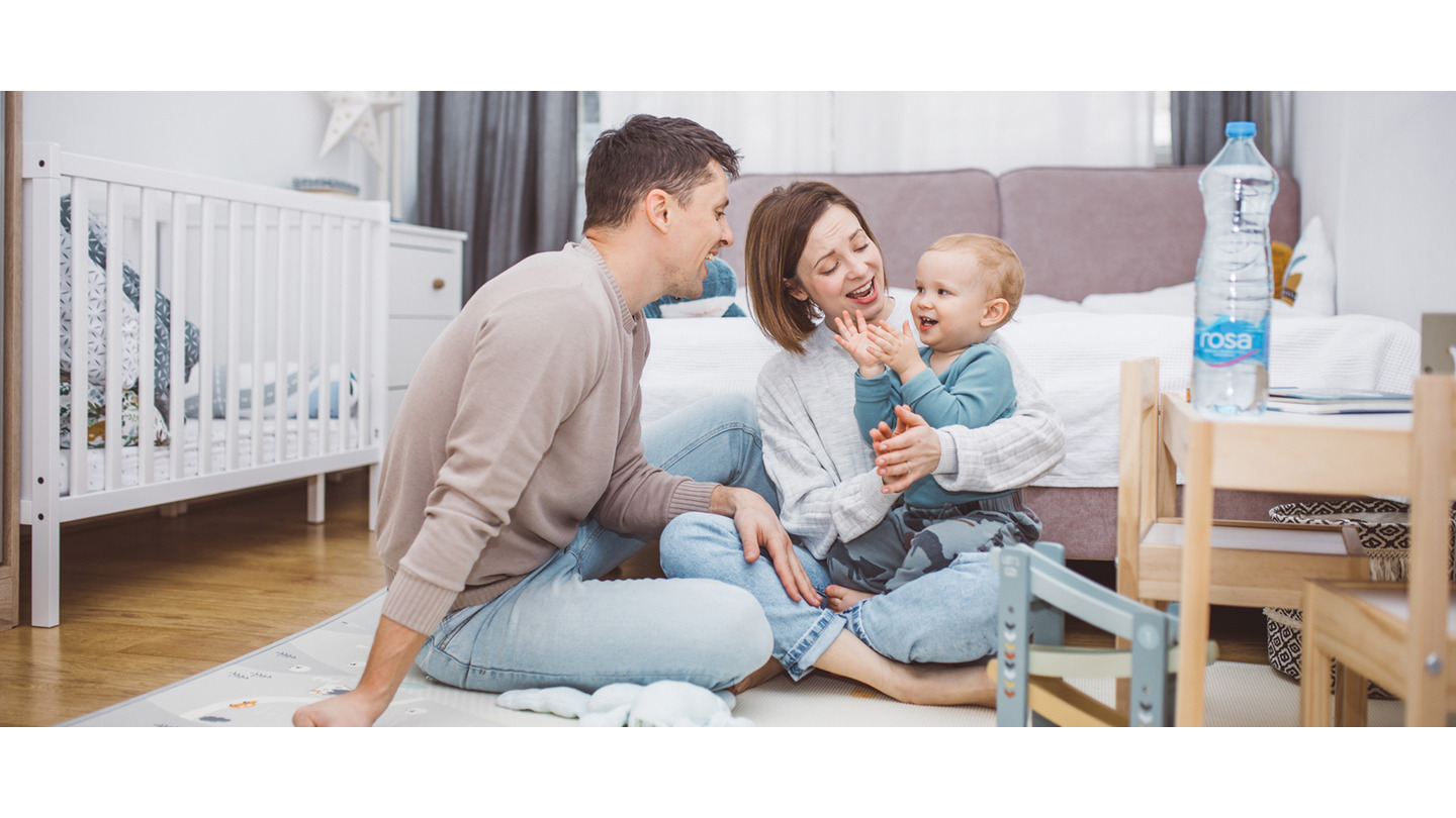 Porodična scena u dečijoj sobi, odrasli sede na podu sa detetom, dok flaša Rosa vode stoji na stolu pored