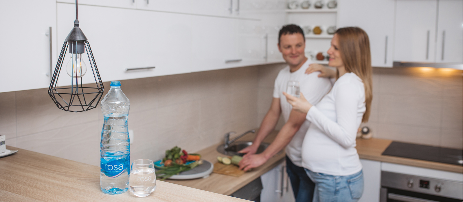 Boca i čaša Rosa vode na kuhinjskom pultu, dok trudnica i muškarac stoje u kuhinji pored isečenog povrća