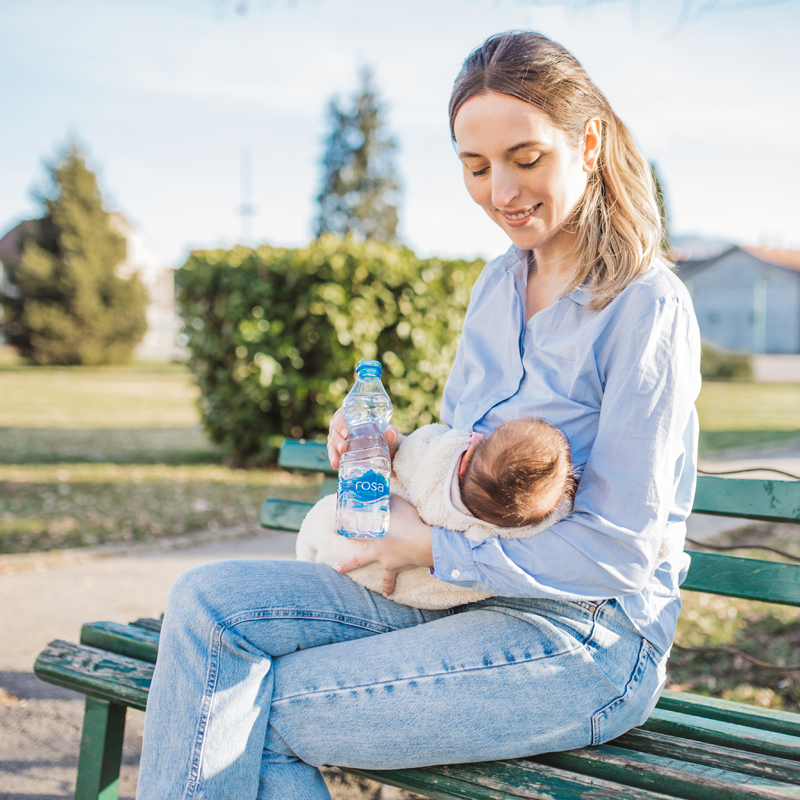 Žena sedi na klupi u parku, drži bebu i bocu Rosa vode