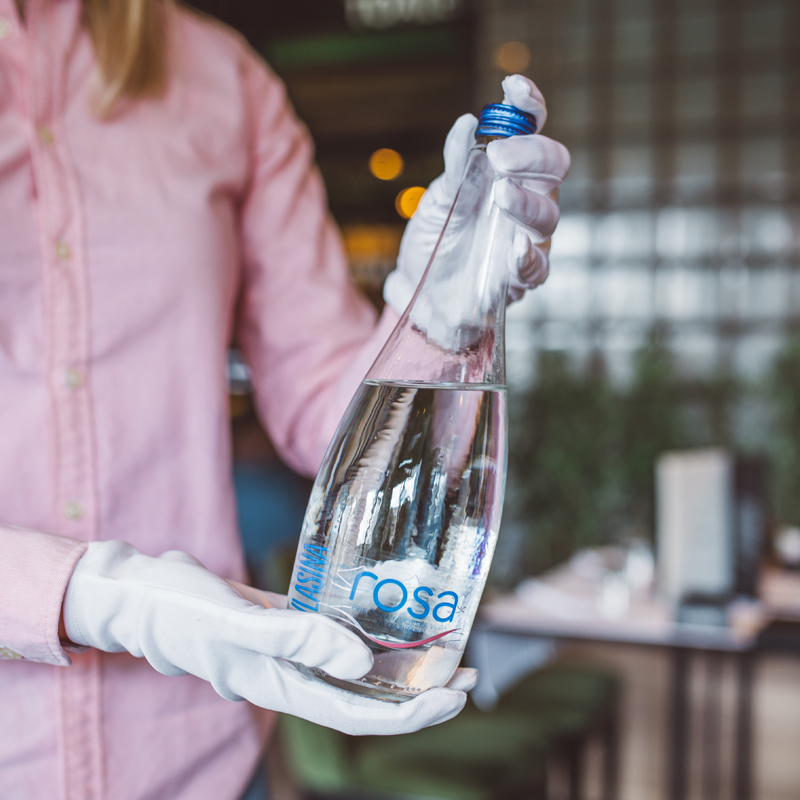 Osoba u ružičastoj košulji i belim rukavicama drži flašu Rosa vode u restoranskom okruženju