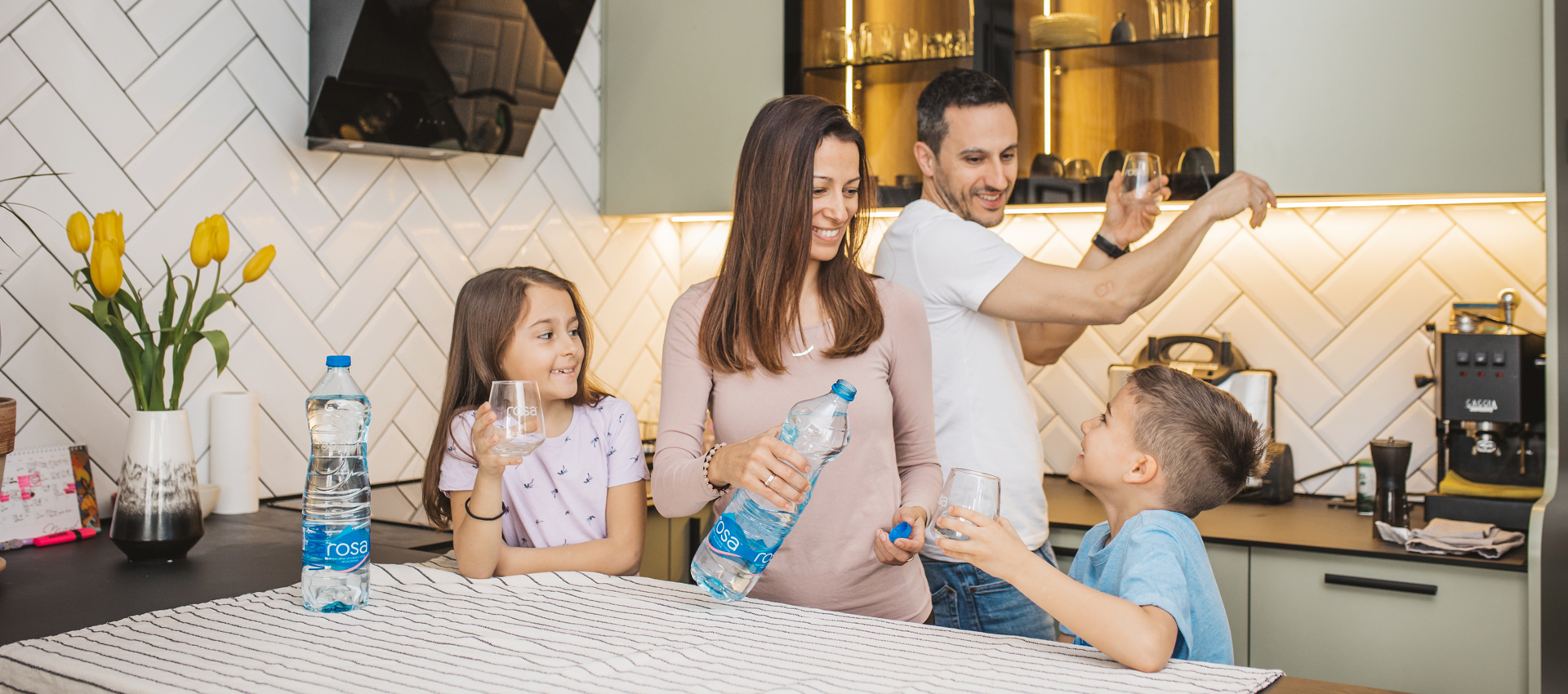 Porodica u kuhinji sipa i pije Rosa vodu iz čaša, sa bocom vode na stolu i žutim tulipanima u pozadini