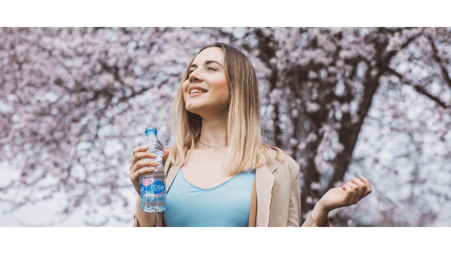 Osoba u plavoj majici i bež sakou drži bocu Rosa vode, stojeći ispred cvetajućih stabala, uz tekst "Rosa Vlasina i zdravlje"