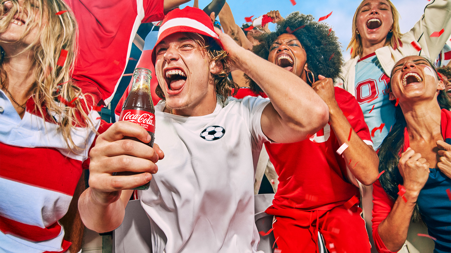 Soccer fan holding a Coca-Cola surrounded by other fans all yelling during a match