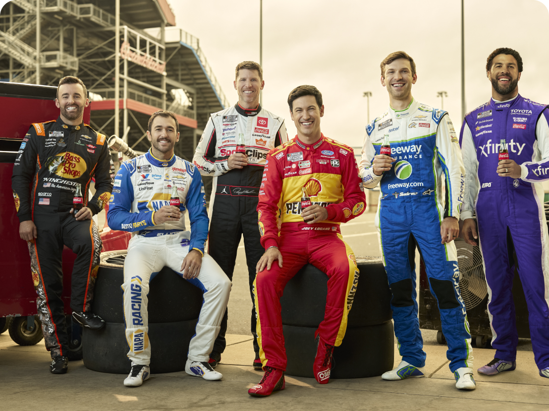 Six NASCAR drivers in racing suits hold Coca‑Cola® bottles while posing together by tires at the DAYTONA 500® track.
