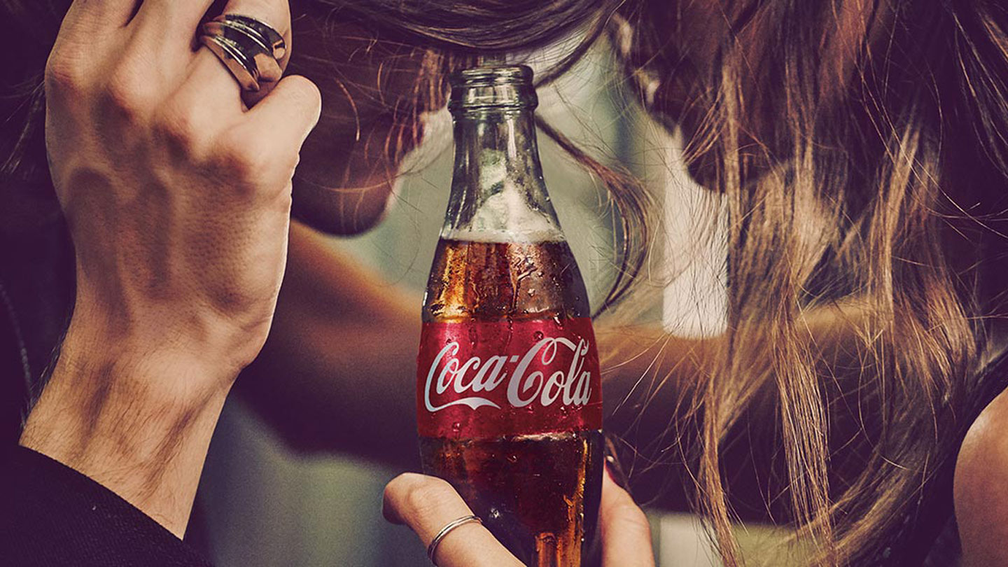 baner sa flašicom Coca-Cole u fokusu i momkom i devojkom sa strana